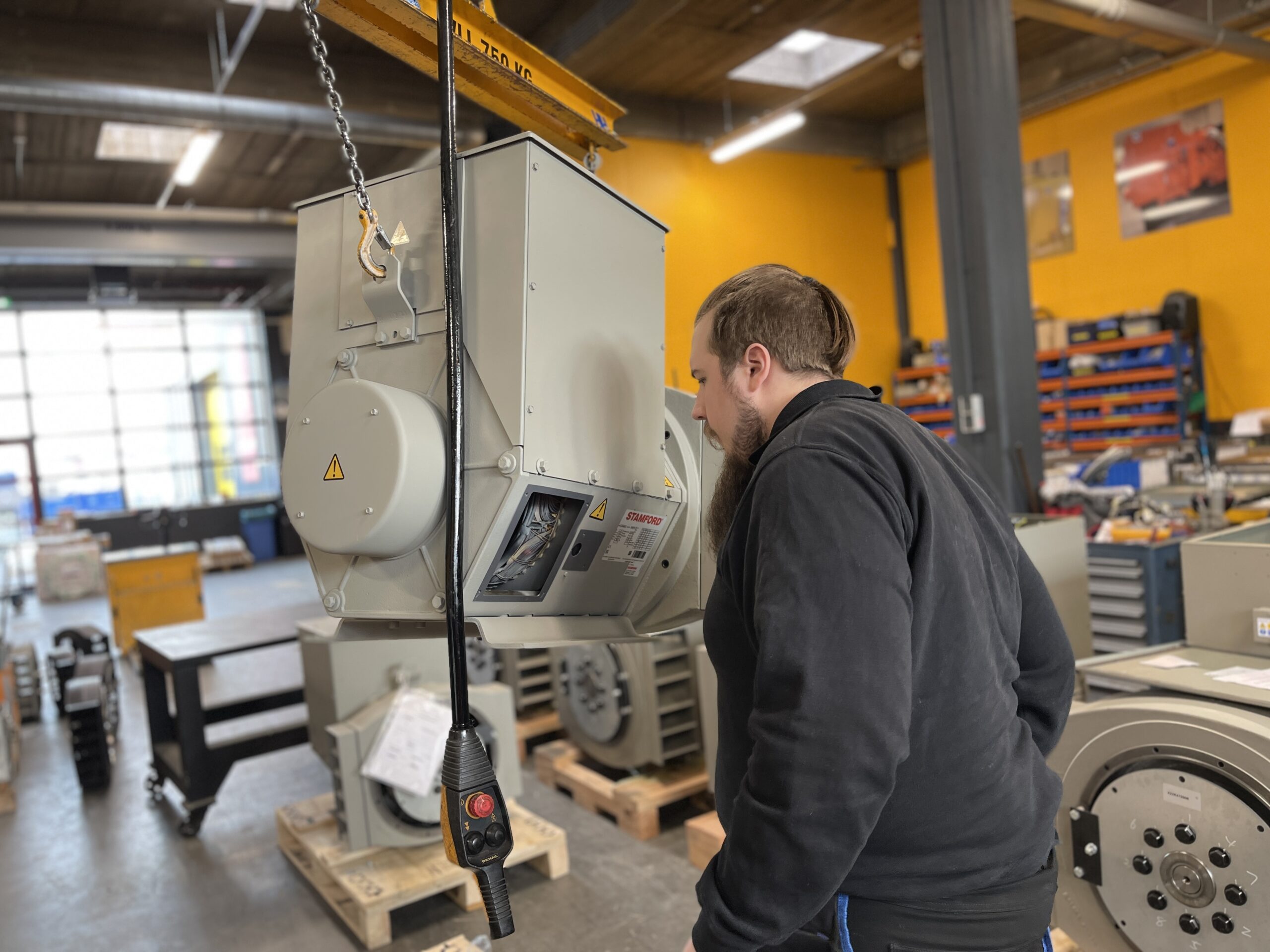 A man inspects a large industrial machine suspended by a yellow overhead hoist in a workshop with yellow walls, organised shelves, and clear signs of organisatie in the background.