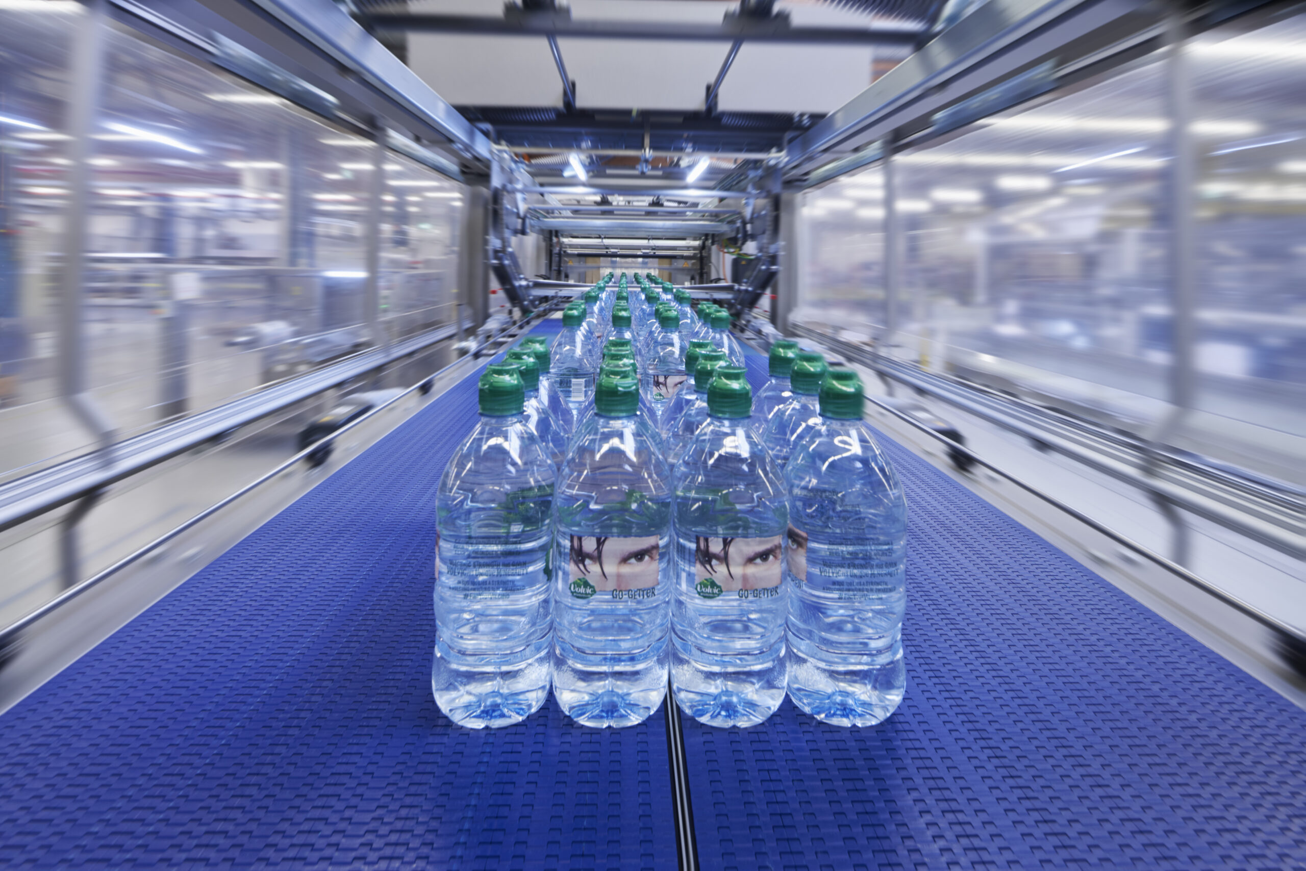 Rijen doorzichtige plastic waterflessen met groene doppen bewegen over een blauwe transportband in een helder verlichte, moderne bottelfabriek. De achtergrond is wazig, waardoor de beweging en snelheid van de productie wordt benadrukt.