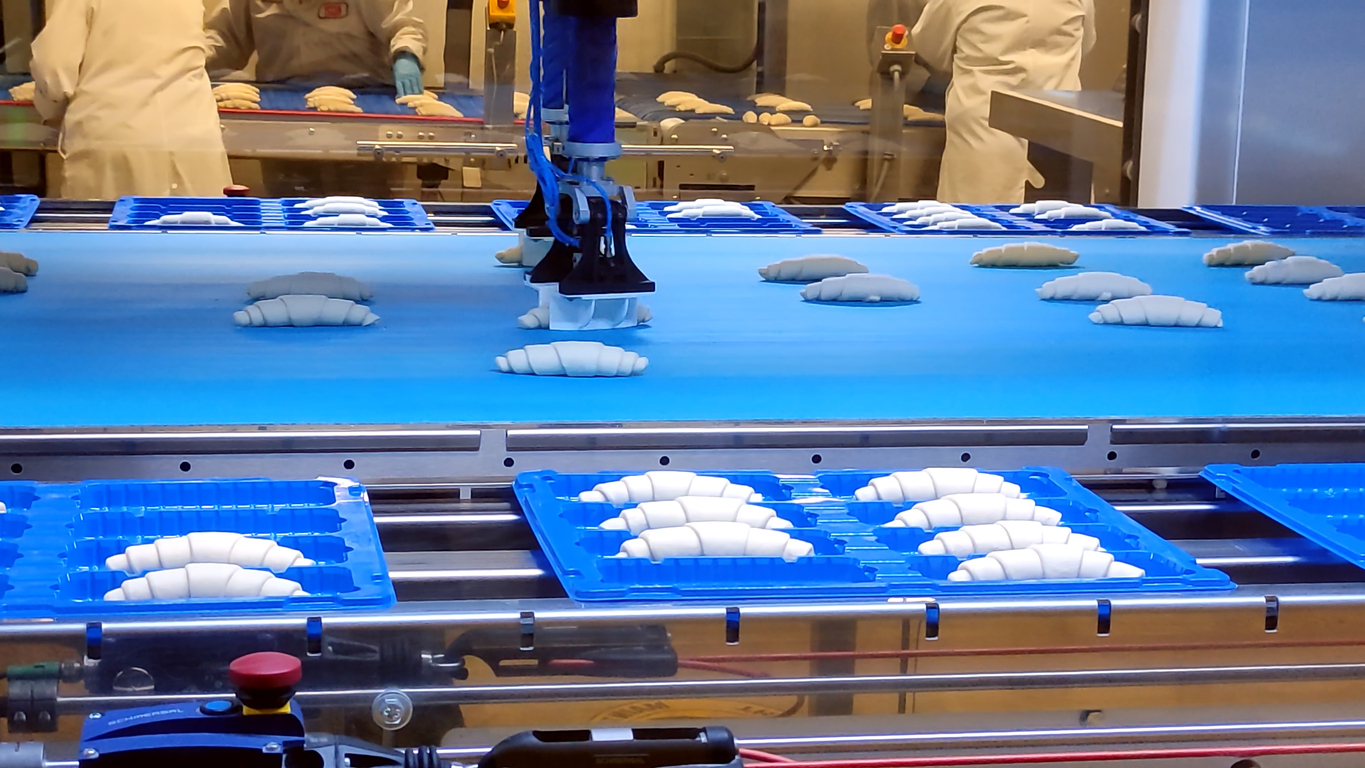 Een robotarm plaatst rauwe croissants op een blauwe transportband in een fabriek, terwijl arbeiders in witte uniformen op de achtergrond werken. Blauwe dienbladen gevuld met croissants zijn zichtbaar op de lopende band.