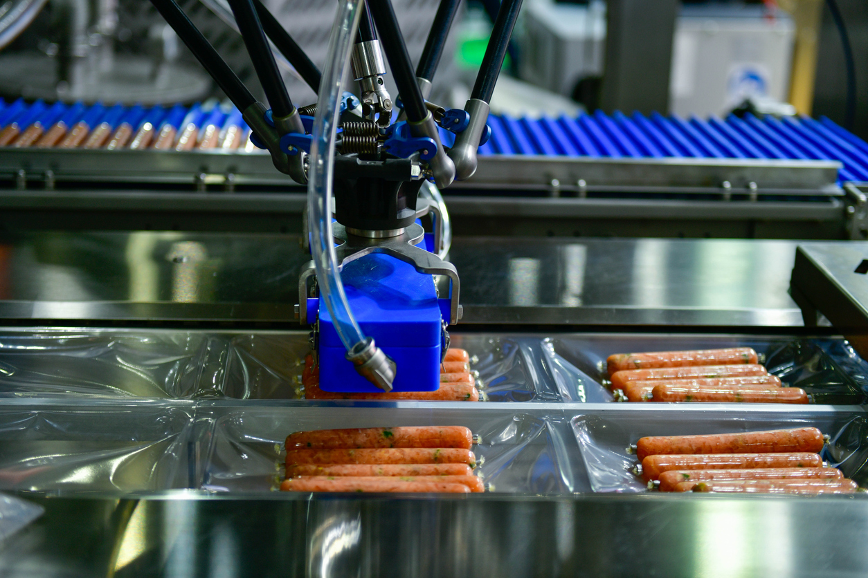 Een robotarm in een voedselverwerkingsfabriek plaatst worsten in doorzichtige plastic bakjes op een metalen transportband en maakt ze klaar om verpakt te worden.