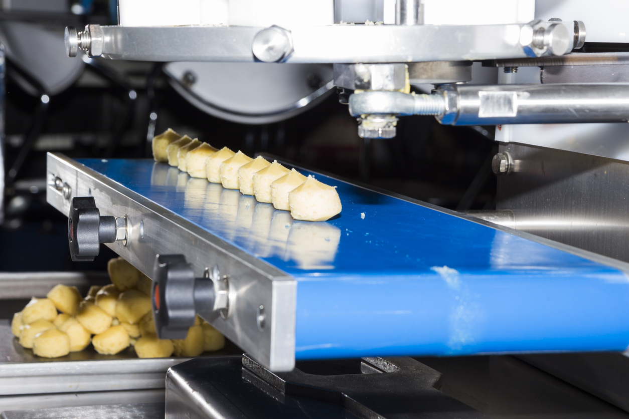 Close-up van een voedselverwerkingsmachine die kleine stukjes deeg vormt en snijdt op een blauwe transportband, met afgewerkte deegstukken verzameld in een bak eronder.