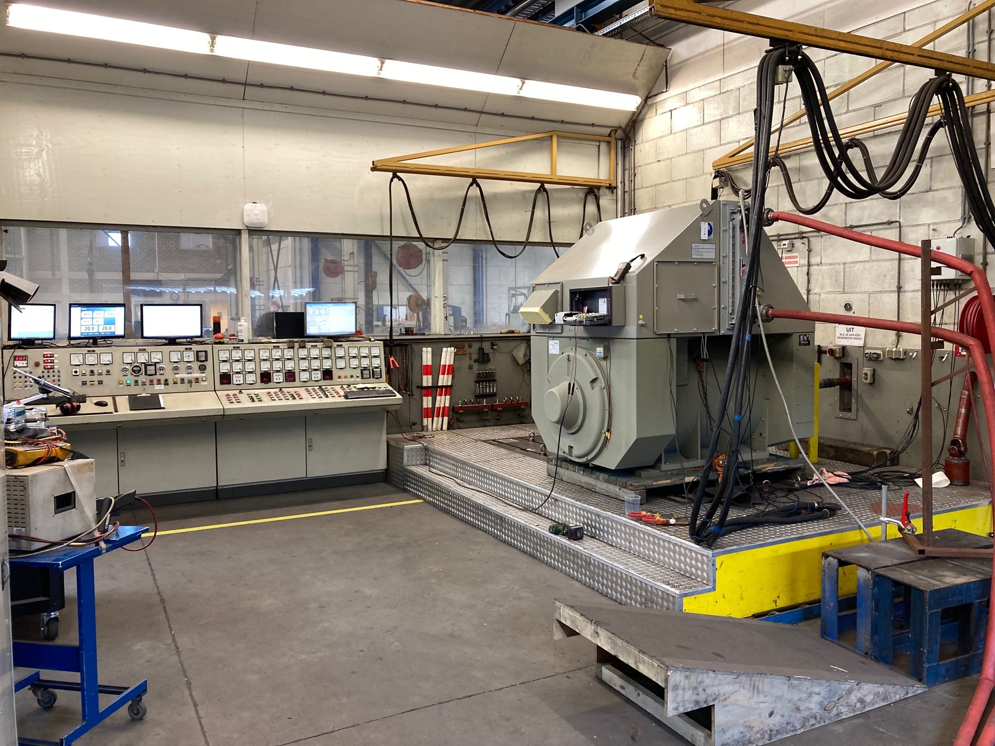 Een grote industriële controlekamer met een paneel vol knoppen, schakelaars en schermen voor de classificatie van generatoren, tegenover een zware machine op een verhoogd platform, omringd door kabels en gereedschappen in een ruime, goed verlichte fabrieksomgeving.