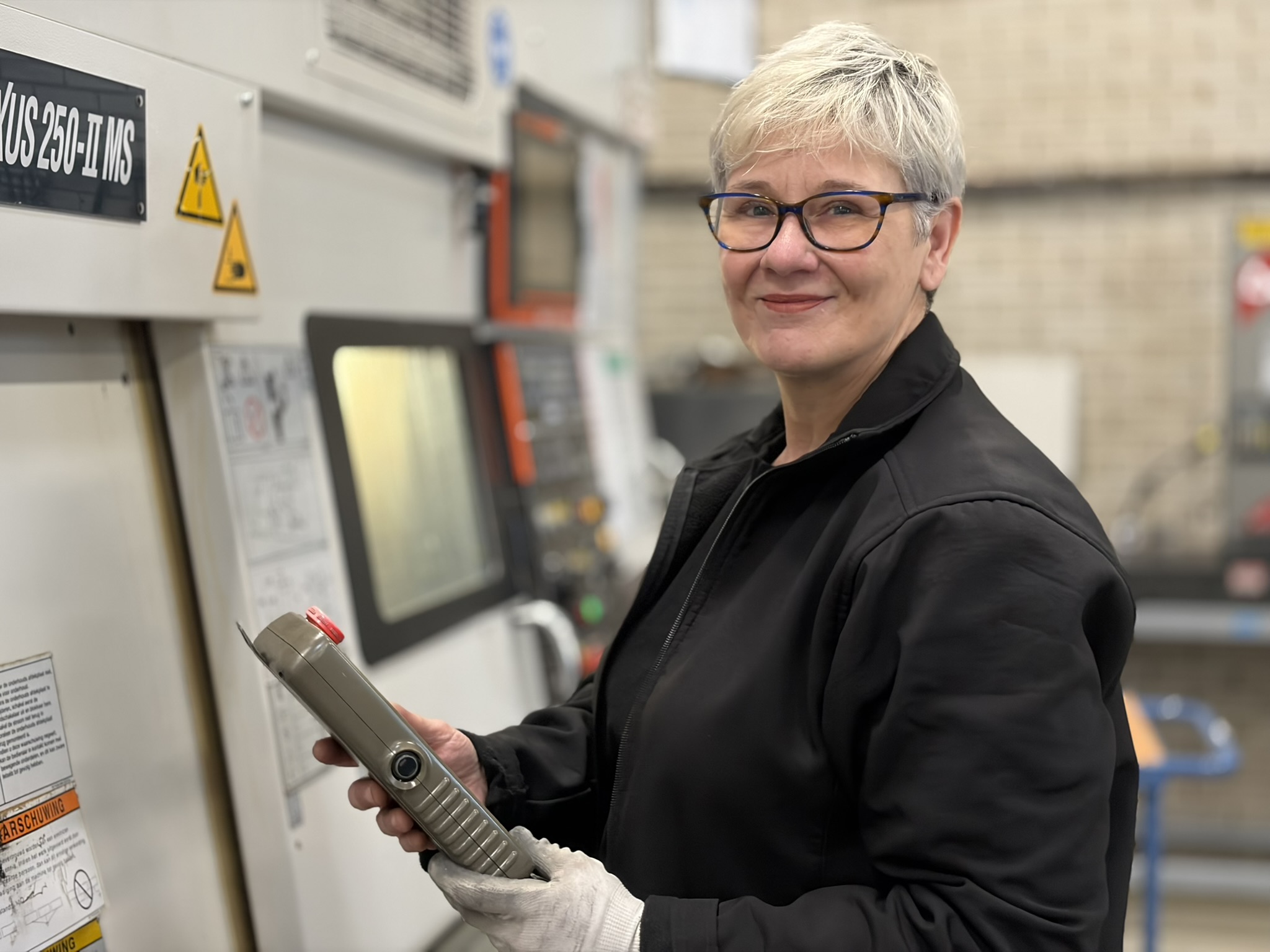 With experience at Caldic Techniek, Jolanda Stadhouders—sporting short gray hair and glasses—stands before industrial machinery, wearing a black jacket and white gloves, holding a control panel and smiling at the camera.