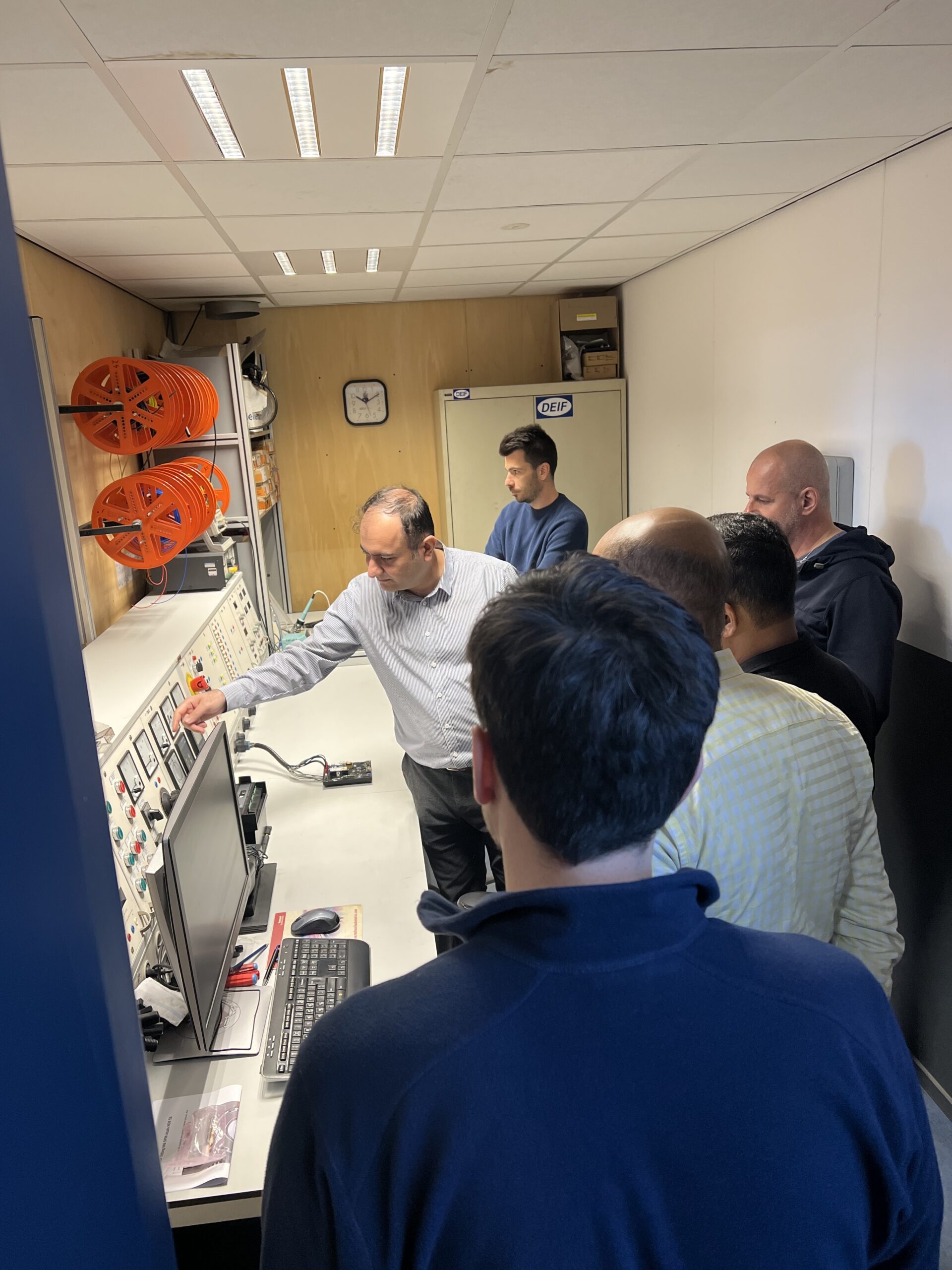 Een groep van zes mannen staat in een kleine controlekamer en één van hen wijst naar een bedieningspaneel aan de muur. Monitoren, toetsenborden en oranje slanghaspels zijn zichtbaar; een klok en een kast staan op de achtergrond.