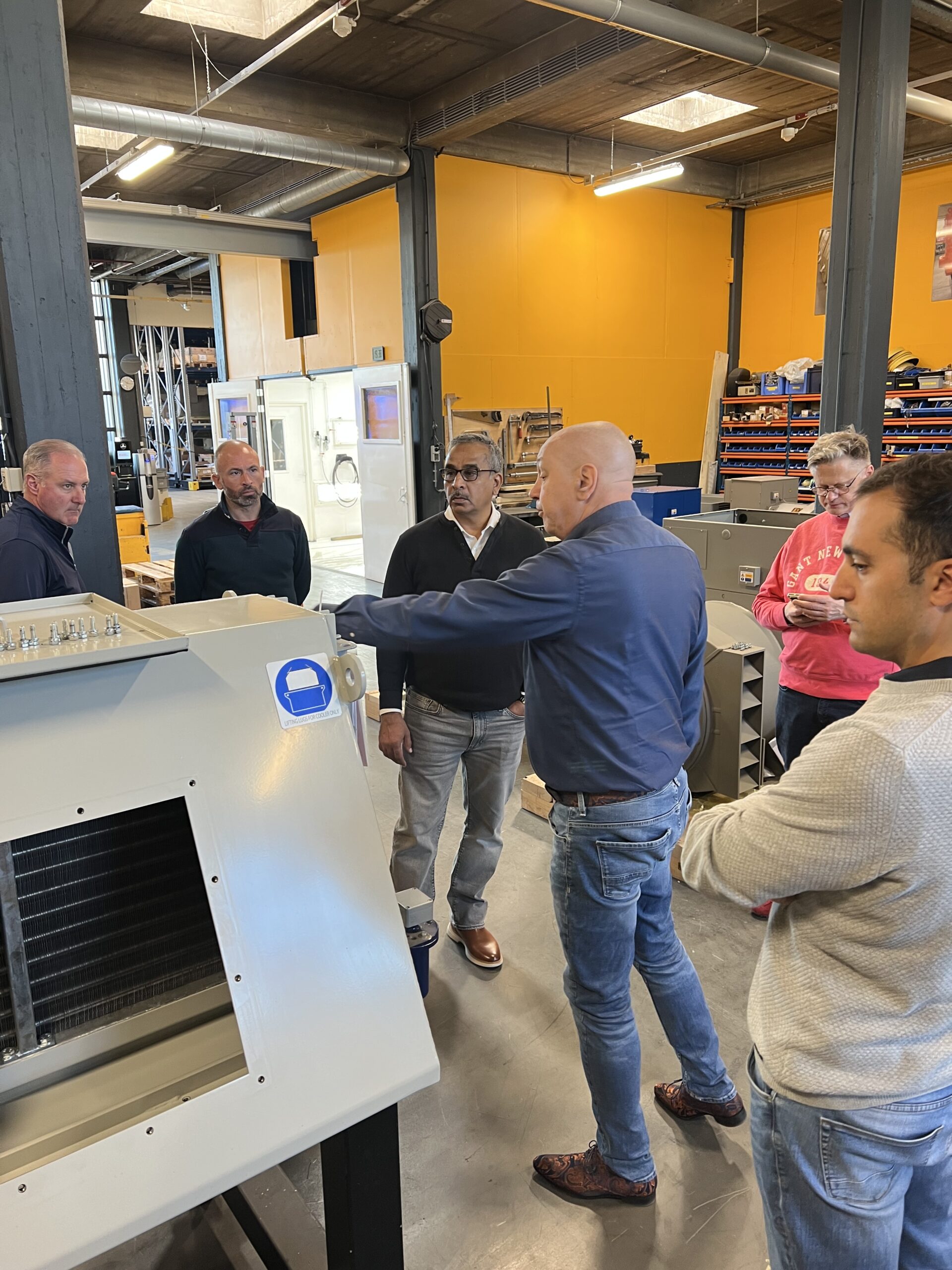 Een groep van zes mensen staat rond een grote industriële machine in een werkplaats en is in gesprek. Eén persoon wijst naar de machine terwijl de anderen aandachtig luisteren. Op de achtergrond zijn planken en apparatuur zichtbaar.