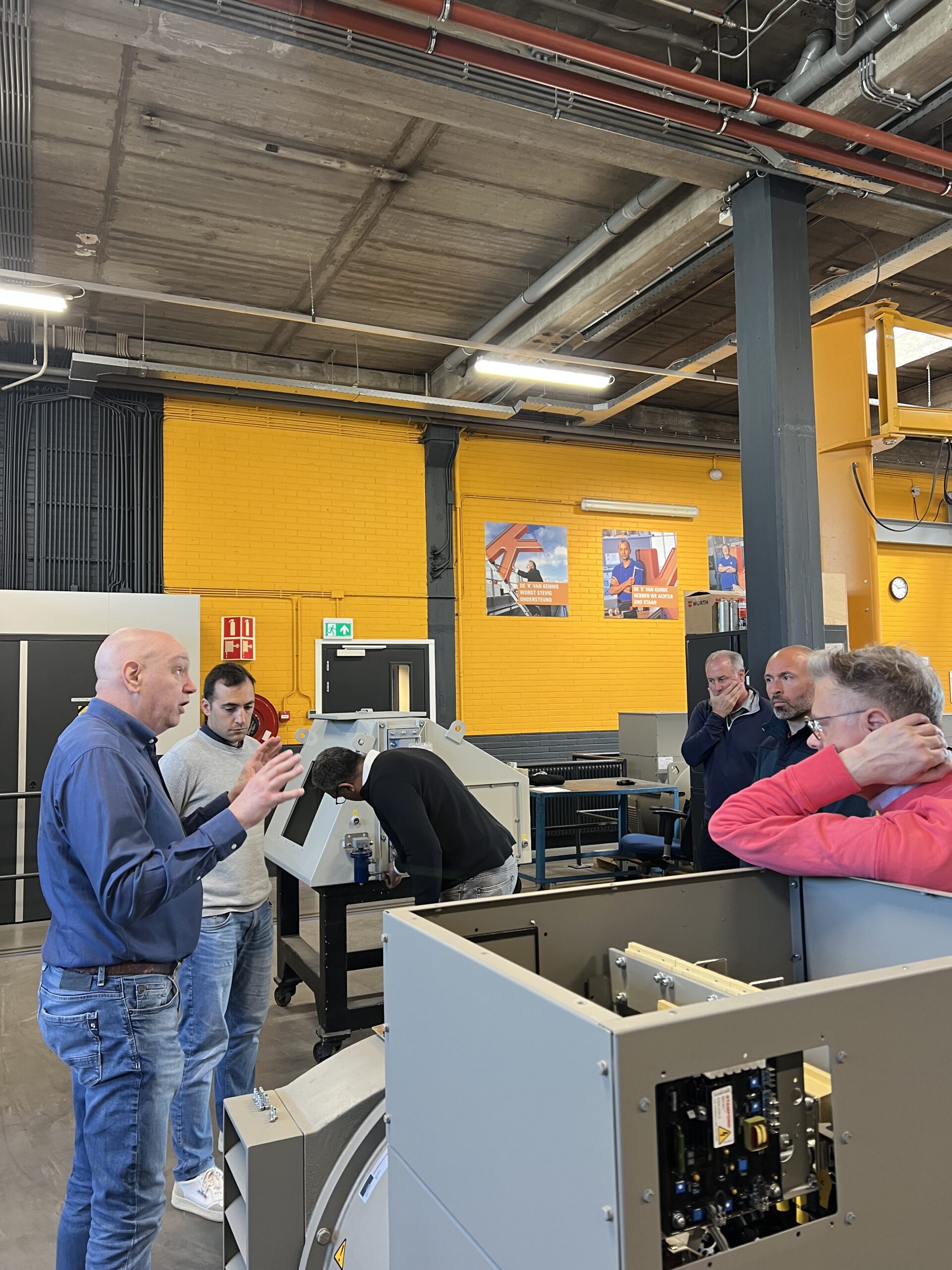 Een groep van zes mensen staat in een industriële werkruimte, in gesprek rond grote machines. Eén man gebaart met zijn handen terwijl de anderen aandachtig luisteren. De ruimte heeft gele bakstenen muren en zichtbare plafondpijpen.