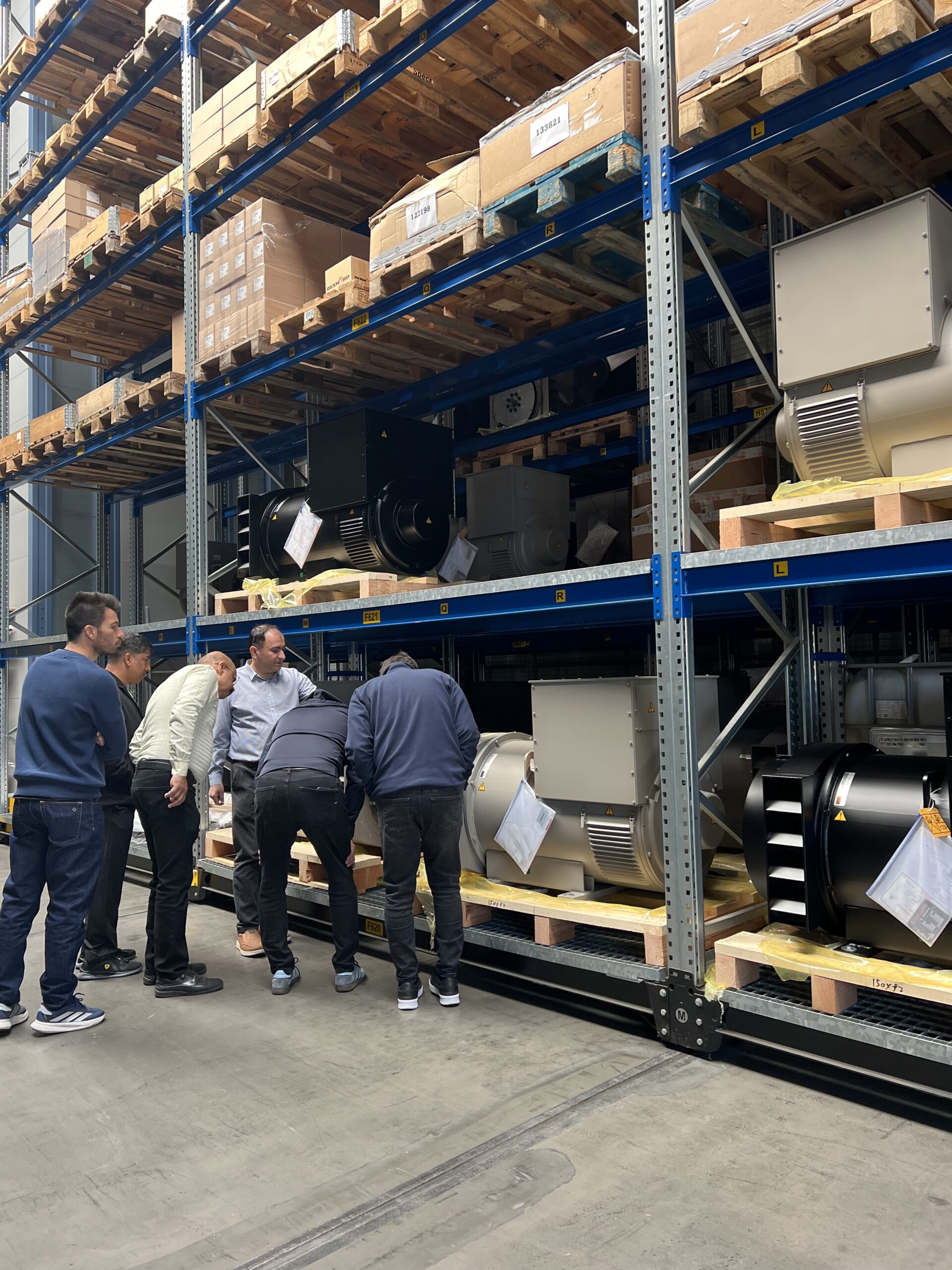 Vijf mannen staan dicht op elkaar en onderzoeken grote industriële apparatuur op lagere magazijnplanken, omringd door houten pallets en metalen rekken volgestapeld met dozen en machines.