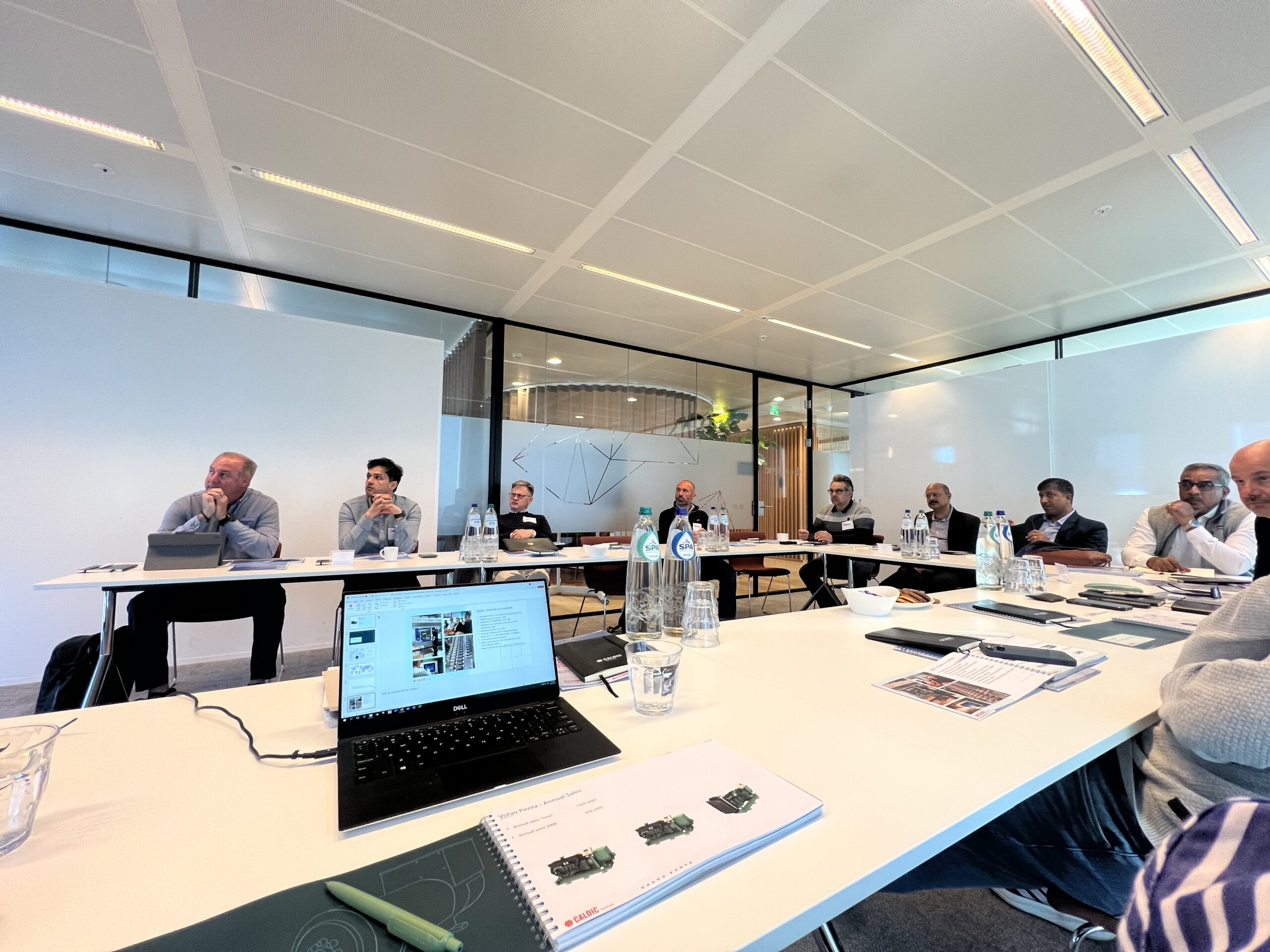 Een groep mensen zit rond een grote vergadertafel in een moderne vergaderruimte, met laptops, waterflessen en documenten voor zich. Op de voorgrond is een laptop te zien met een webpagina.